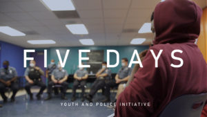 Title card from video showing teenager in a hooded sweatshirt, hood up and front pulled over his face. Across the room sit seven police officers Text overlay reads "Five Days: Youth and Police Initiative."