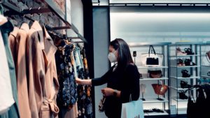 Woman wearing COVID mask and holding a shopping bag looks through rack of clothes in store